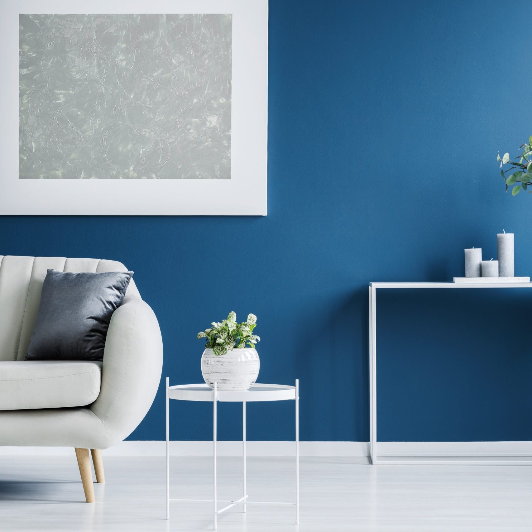 Metal console table with plant in vase and candles standing against blue wall in living room interior with light grey sofa and modern poster